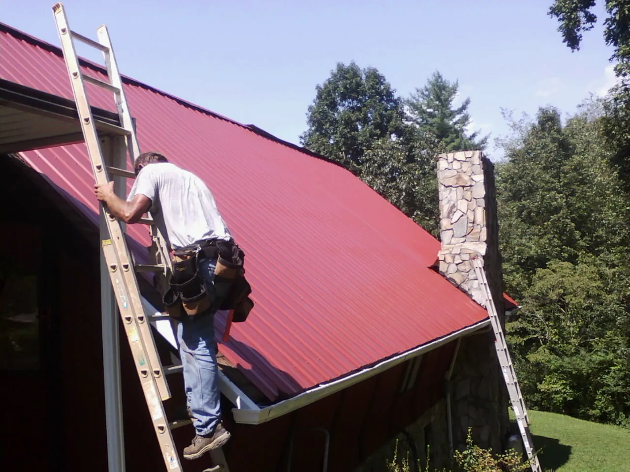 Roofer on a ladder beside a red metal roof and stone chimney — Soffit & Fascia Repair by Roofing Brothers in Comfrey